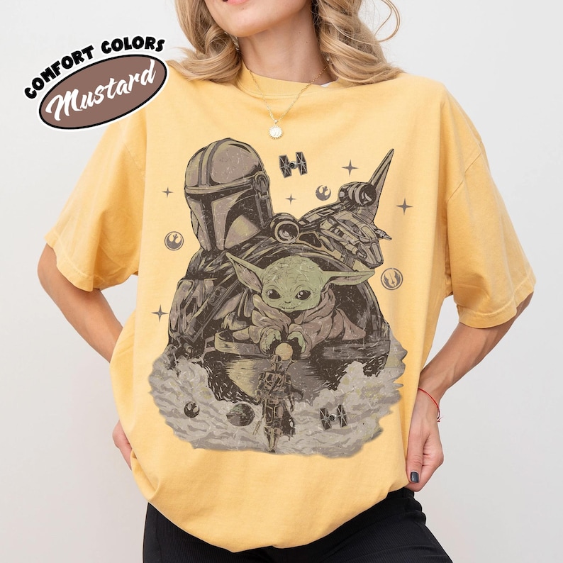 Puede incluir: Camiseta color mostaza con un gr&aacute;fico de The Mandalorian, Grogu y una nave espacial en tonos grises y marrones. El texto "COMFORT COLORS" y "Mustard" est&aacute; impreso en la esquina superior izquierda.