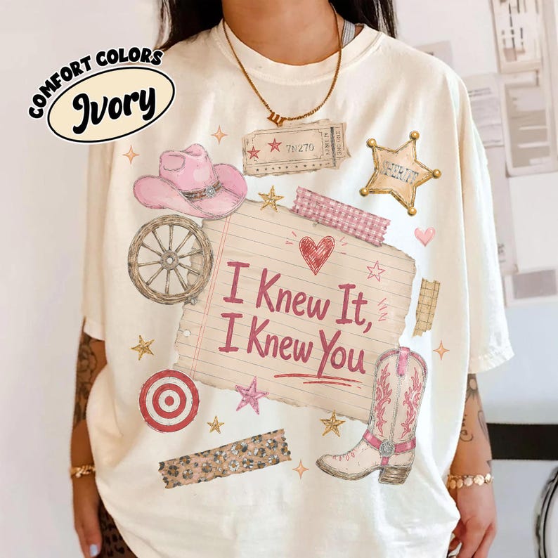 Puede incluir: Una camiseta color marfil con un estampado gr&aacute;fico que incluye un sombrero de vaquero rosa, una rueda de carreta, una estrella de sheriff, una bota vaquera y una hoja de papel rayado con el texto I Knew It, I Knew You, rodeado de estrellas.