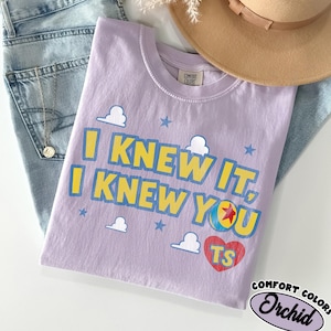 May include: A light purple short-sleeve t-shirt featuring the yellow text I KNEW IT, I KNEW YOU with blue outlines. The design includes white cloud shapes, blue stars, a colorful ball with a red star, and a red heart containing the yellow letters TS.