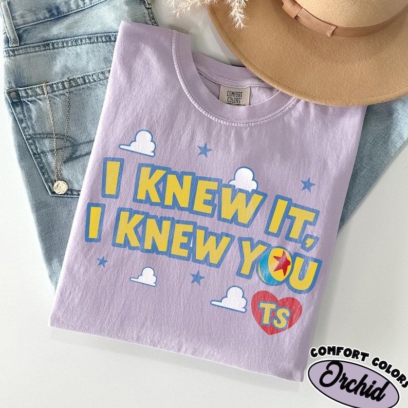 May include: A light purple short-sleeve t-shirt featuring the yellow text I KNEW IT, I KNEW YOU with blue outlines. The design includes white cloud shapes, blue stars, a colorful ball with a red star, and a red heart containing the yellow letters TS.