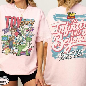 Pode incluir: Uma t-shirt rosa claro com gr&aacute;ficos de Toy Story. A frente mostra Woody, Buzz Lightyear, Rex e outras personagens com o texto "Toy Story" e "Since 1995". As costas apresentam o texto "To Infinity and Beyond" e "Est. 1995".