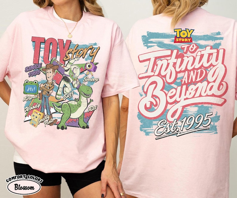 Pode incluir: Uma t-shirt rosa claro com gr&aacute;ficos de Toy Story. A frente mostra Woody, Buzz Lightyear, Rex e outras personagens com o texto "Toy Story" e "Since 1995". As costas apresentam o texto "To Infinity and Beyond" e "Est. 1995".