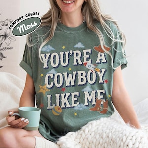 May include: A moss green t-shirt featuring the text "YOU'RE A COWBOY LIKE ME" in a distressed font. The graphic includes illustrations of a rocket, a sheriff star, a ball, and cowboy boots, surrounded by small clouds and stars in various colors.