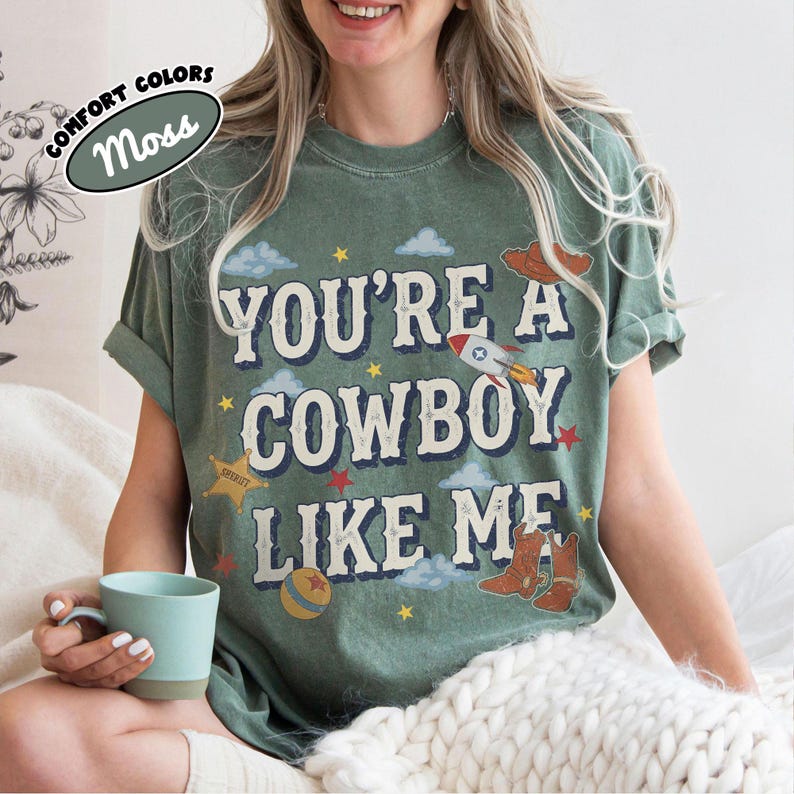 May include: A moss green t-shirt featuring the text "YOU'RE A COWBOY LIKE ME" in a distressed font. The graphic includes illustrations of a rocket, a sheriff star, a ball, and cowboy boots, surrounded by small clouds and stars in various colors.