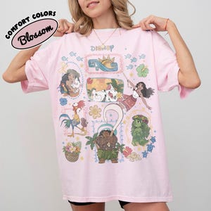 May include: A pale pink t-shirt with Disney character illustrations. The shirt displays the word "Disney" and the text "Comfort Colors Blossom" in a circular design. The graphics include cartoon characters and floral patterns.