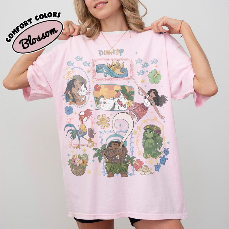 May include: A pale pink t-shirt with Disney character illustrations. The shirt displays the word "Disney" and the text "Comfort Colors Blossom" in a circular design. The graphics include cartoon characters and floral patterns.