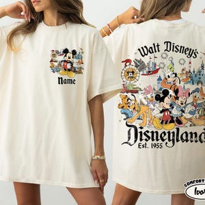 Peut inclure: T-shirt ivoire avec des motifs de Mickey Mouse et Disneyland. L'avant pr&eacute;sente Mickey avec le texte "Name". Le dos affiche des personnages Disney, un ch&acirc;teau et le texte "Walt Disney's Disneyland Est. 1955".