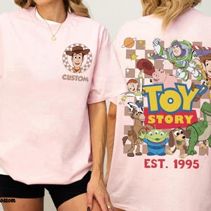 Puede incluir: Camiseta rosa claro con un gr&aacute;fico frontal de Woody de Toy Story y la palabra "CUSTOM". La parte trasera presenta personajes de Toy Story y el texto "TOY STORY EST. 1995".