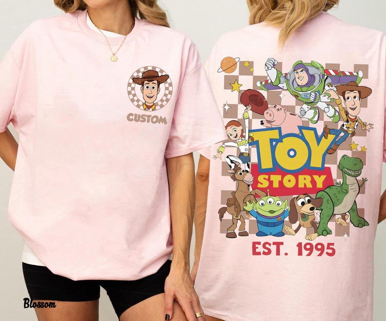 Puede incluir: Camiseta rosa claro con un gr&aacute;fico frontal de Woody de Toy Story y la palabra "CUSTOM". La parte trasera presenta personajes de Toy Story y el texto "TOY STORY EST. 1995".