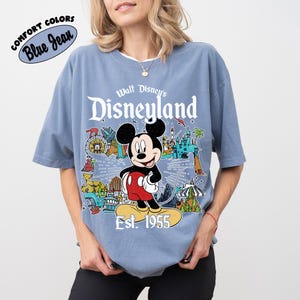 Op de afbeelding: Een blauw T-shirt met korte mouwen en een afbeelding van Mickey Mouse boven de tekst "Walt Disney's Disneyland Est. 1955". Het ontwerp bevat kleurrijke illustraties van attracties in het park, waaronder een kasteel, een trein en een reuzenrad.