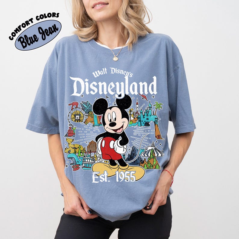 Op de afbeelding: Een blauw T-shirt met korte mouwen en een afbeelding van Mickey Mouse boven de tekst "Walt Disney's Disneyland Est. 1955". Het ontwerp bevat kleurrijke illustraties van attracties in het park, waaronder een kasteel, een trein en een reuzenrad.
