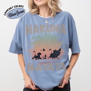 May include: A blue t-shirt with the words "HAKUNA MATATA" in a distressed font. The shirt features a graphic of silhouettes of characters from The Lion King, with a colorful gradient background. The shirt has the words "Comfort Colors Blue Jean" on the upper left.