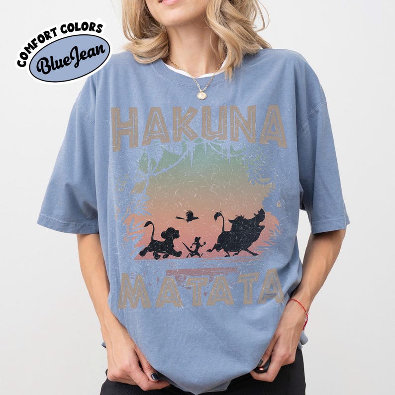 May include: A blue t-shirt with the words "HAKUNA MATATA" in a distressed font. The shirt features a graphic of silhouettes of characters from The Lion King, with a colorful gradient background. The shirt has the words "Comfort Colors Blue Jean" on the upper left.