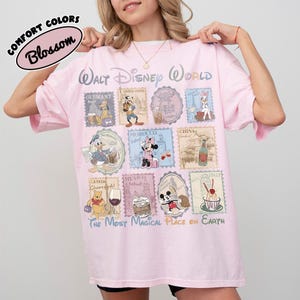 May include: A light pink t-shirt with a Walt Disney World design. The design features various Disney characters and stamps from different countries. The text "COMFORT COLORS Blossom" is at the top left. The phrase "THE MOST MAGICAL PLACE ON EARTH" is at the bottom.
