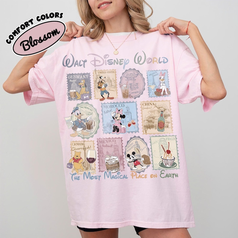 May include: A light pink t-shirt with a Walt Disney World design. The design features various Disney characters and stamps from different countries. The text "COMFORT COLORS Blossom" is at the top left. The phrase "THE MOST MAGICAL PLACE ON EARTH" is at the bottom.