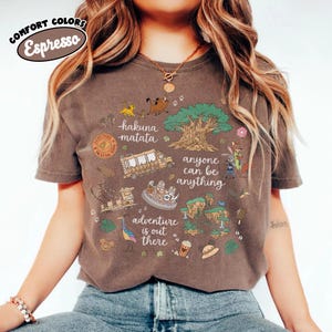 Puede incluir: Camiseta color espresso con un dise&ntilde;o gr&aacute;fico colorido con las palabras "Hakuna Matata", "Anyone Can Be Anything" y "Adventure is Out There". El dise&ntilde;o incluye im&aacute;genes de dibujos animados de animales, un &aacute;rbol y un autob&uacute;s.