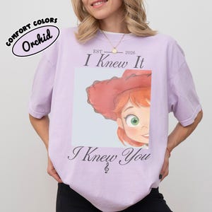 Op de afbeelding: Een lichtpaars t-shirt met een afbeelding van een stripfiguur met rood haar en een rode hoed. De tekst luidt "EST 2026 I Knew It I Knew You" met een kleine muzieknoot. Een "COMFORT COLORS Orchid" label is zichtbaar in de linkerbovenhoek.