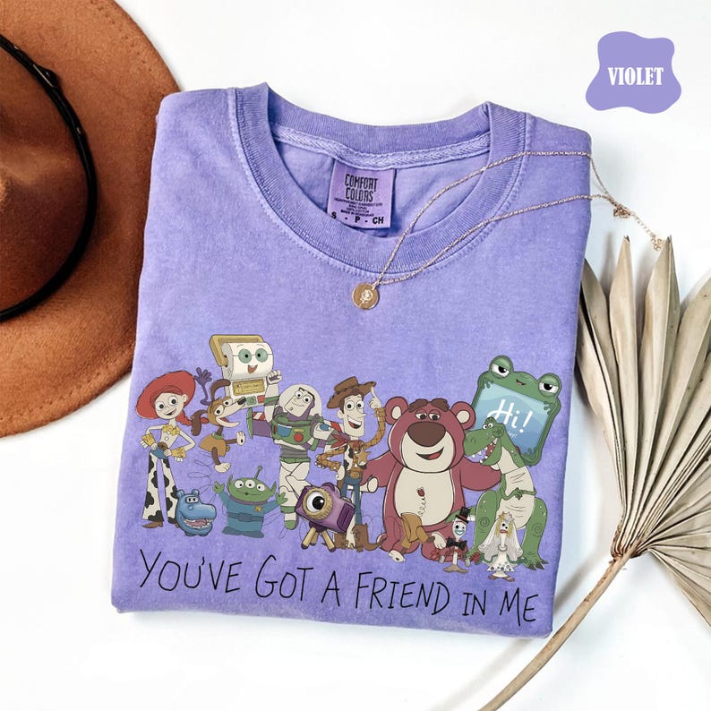May include: A violet short-sleeve t-shirt featuring a graphic print of various animated characters from the Toy Story franchise. The text "YOU'VE GOT A FRIEND IN ME" is printed in black below the characters. The shirt has a crew neckline.