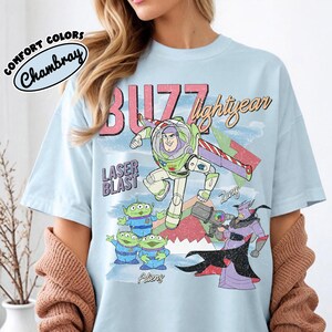 May include: Pale blue t-shirt with a graphic of Buzz Lightyear, aliens, and Zurg from Toy Story. The shirt has the words "BUZZ lightyear" and "LASER BLAST" printed on it. The shirt is made of chambray material.