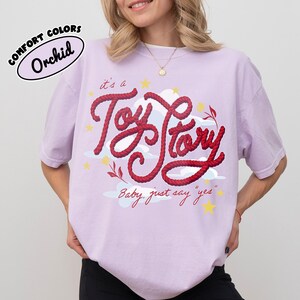 May include: A light purple t-shirt featuring a graphic design with the red rope-style text "it's a Toy Story" and "Baby, just say "yes"" surrounded by white clouds and yellow stars. The top left corner includes a "COMFORT COLORS Orchid" label.