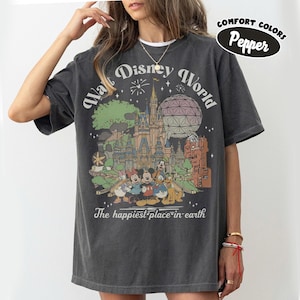 May include: Dark gray t-shirt featuring a colorful graphic of Walt Disney World with Mickey Mouse, Minnie Mouse, Donald Duck, and Goofy. The text "Walt Disney World" and "The happiest place on earth" are printed on the shirt. The shirt is labeled "Comfort Colors Pepper".