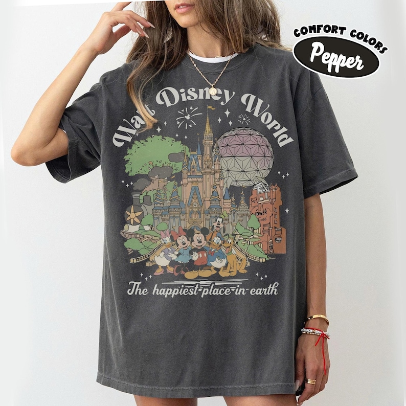 May include: Dark gray t-shirt featuring a colorful graphic of Walt Disney World with Mickey Mouse, Minnie Mouse, Donald Duck, and Goofy. The text "Walt Disney World" and "The happiest place on earth" are printed on the shirt. The shirt is labeled "Comfort Colors Pepper".