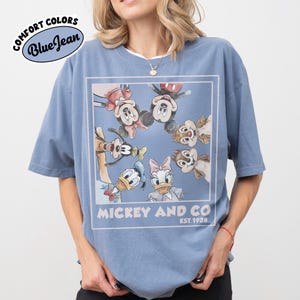 Puede incluir: Camiseta azul con un gr&aacute;fico de Mickey Mouse, Minnie Mouse, Pato Donald, Daisy, Chip y Chop. La camiseta tiene el texto "Mickey and Co" y "Est 1928". La camiseta est&aacute; etiquetada como "Comfort Colors Blue Jean".