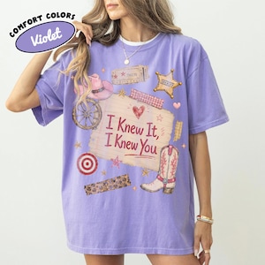 Puede incluir: Una camiseta violeta de manga corta con un estampado gr&aacute;fico de una nota de papel rayado con el texto "I Knew It, I Knew You" rodeado de ilustraciones de un sombrero de vaquero rosa, una estrella de sheriff, una rueda de carreta y una bota.