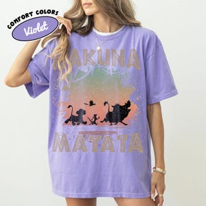 May include: A lilac Comfort Colors t-shirt with the words "Hakuna Matata" and a silhouette of characters from The Lion King. The shirt has a distressed design with a colourful gradient background.