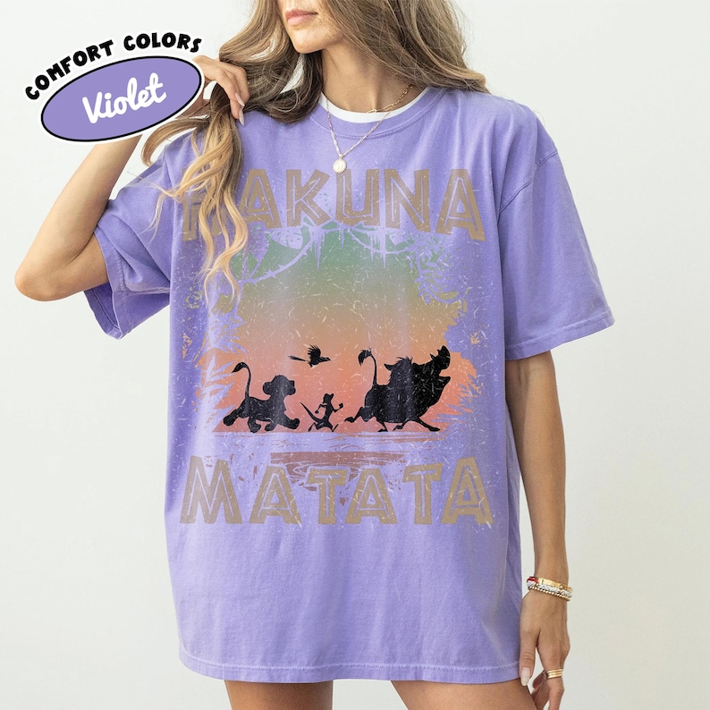 May include: A lilac Comfort Colors t-shirt with the words "Hakuna Matata" and a silhouette of characters from The Lion King. The shirt has a distressed design with a colourful gradient background.