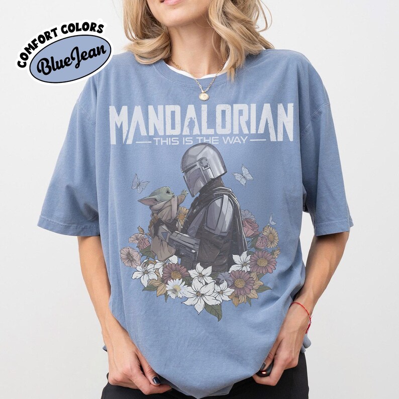 May include: Blue t-shirt with the text "MANDALORIAN - THIS IS THE WAY" and a graphic of the Mandalorian and Grogu surrounded by flowers and butterflies. The shirt has the Comfort Colors and Blue Jean logos.
