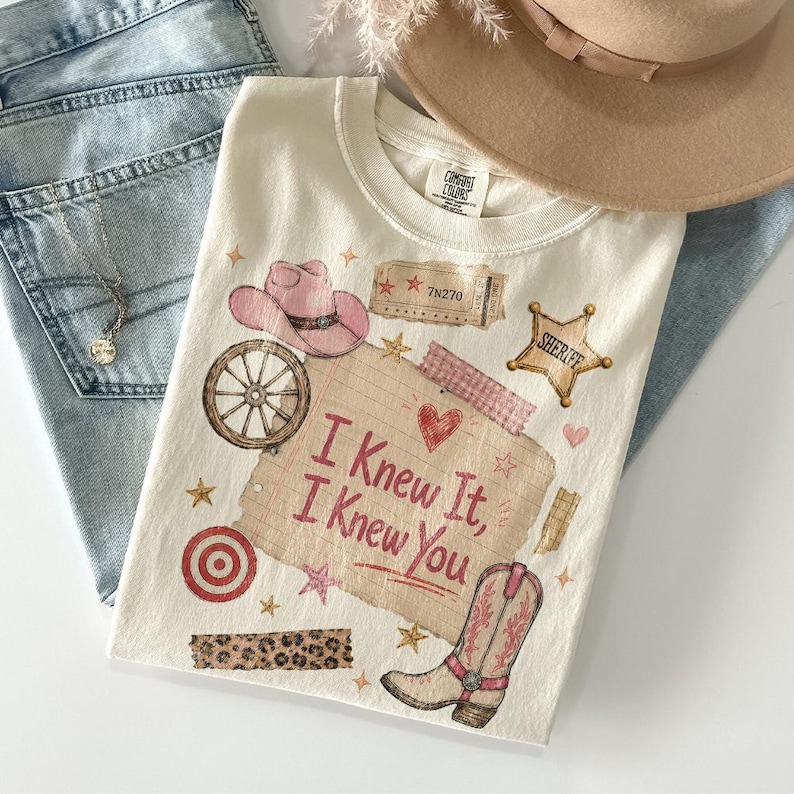 Puede incluir: Una camiseta color crema con un estampado gr&aacute;fico que incluye el texto I Knew It, I Knew You, un sombrero de vaquero rosa, una rueda de carreta, una estrella de sheriff, una bota vaquera, una diana, una franja de leopardo y un ticket 7N270.