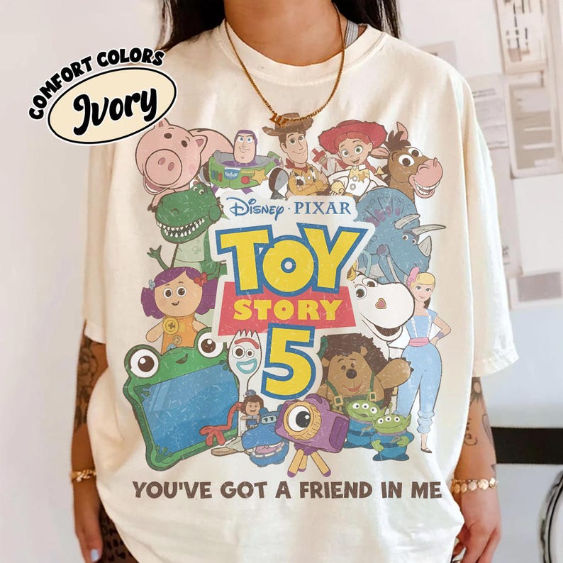 Op de afbeelding: Een ivoorkleurig t-shirt met een grafische print van Disney Pixar Toy Story-personages, waaronder Woody, Buzz Lightyear en Jessie. De tekst luidt Disney Pixar Toy Story 5 en You've Got A Friend In Me. Een Comfort Colors Ivory-label is zichtbaar.