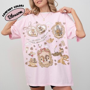May include: A light pink t-shirt with a graphic design featuring Chip and Dale characters in ornate frames and decorative elements. The shirt has the words "COMFORT COLORS" and "Blossom" printed on it.
