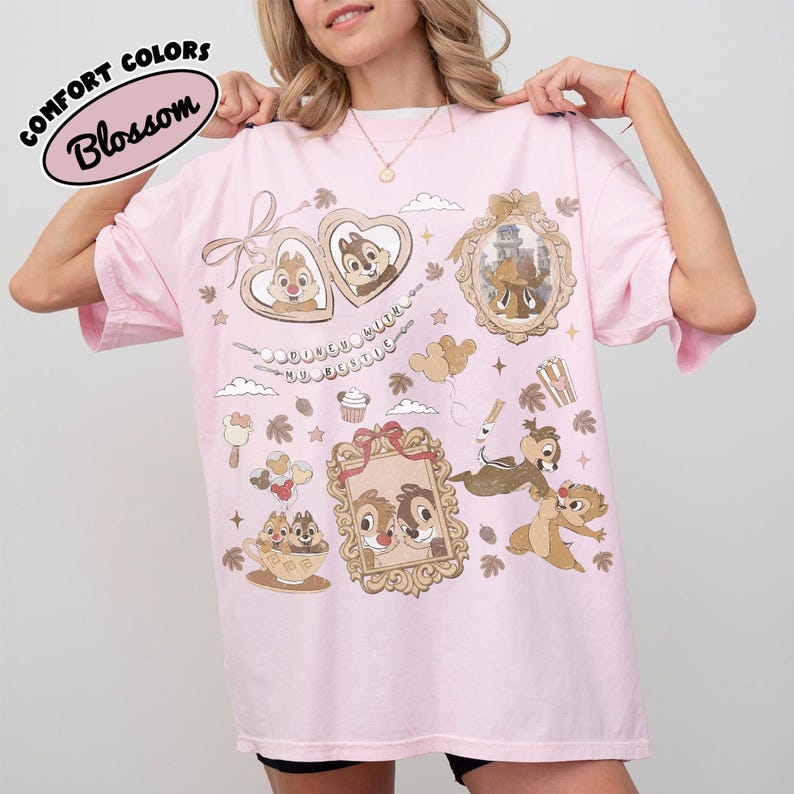May include: A light pink t-shirt with a graphic design featuring Chip and Dale characters in ornate frames and decorative elements. The shirt has the words "COMFORT COLORS" and "Blossom" printed on it.