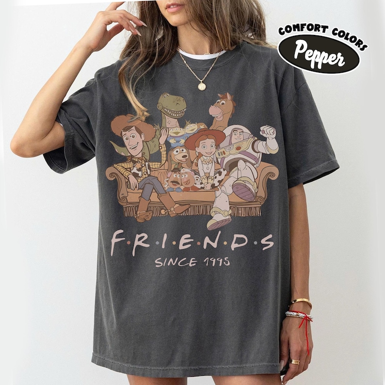 May include: Dark gray t-shirt featuring a graphic of Toy Story characters in a Friends-themed design. The shirt includes the text "FRIENDS" and "SINCE 1995". The shirt has a Comfort Colors Pepper label.