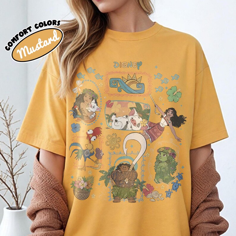 May include: Mustard-coloured t-shirt featuring Disney characters from the movie Moana. The shirt has a crew neck and short sleeves. The text "COMFORT COLORS" and "Mustard" are printed on the upper left. The Disney logo is printed on the upper centre.