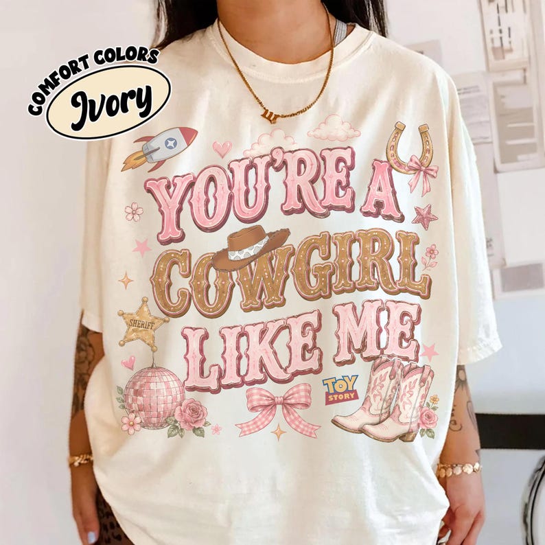 Puede incluir: Una camiseta color marfil con el texto YOU'RE A COWGIRL LIKE ME en letras rosas y marrones. El dise&ntilde;o incluye un cohete, una herradura, un sombrero de vaquero, una estrella de sheriff, una bola de discoteca, un lazo rosa, flores y botas.