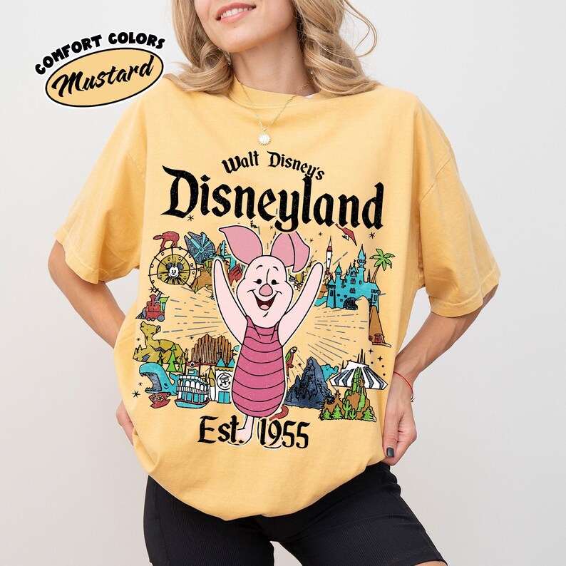 Op de afbeelding: Een mosterdgeel t-shirt met korte mouwen en een afbeelding van Knorretje uit Winnie de Poeh, gecentreerd boven de tekst Walt Disney's Disneyland Est. 1955. Het ontwerp bevat kleurrijke illustraties van diverse attracties en iconen van het park.