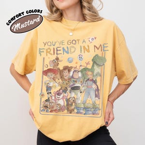 May include: A mustard yellow short-sleeve t-shirt featuring a graphic print of various animated characters from the Toy Story franchise. The text "YOU'VE GOT A FRIEND IN ME" is printed above the characters in a distressed, multi-colored font.