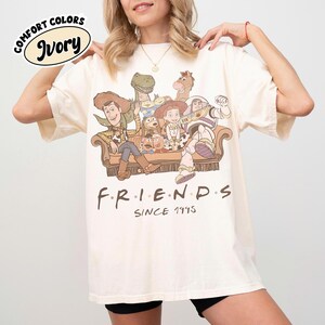May include: Ivory-colored t-shirt featuring a graphic of characters from the movie Toy Story, arranged like the TV show Friends. The text "F.R.I.E.N.D.S" and "Since 1995" are printed below the graphic. The shirt has the words "Comfort Colors" and "Ivory" printed on the top left.