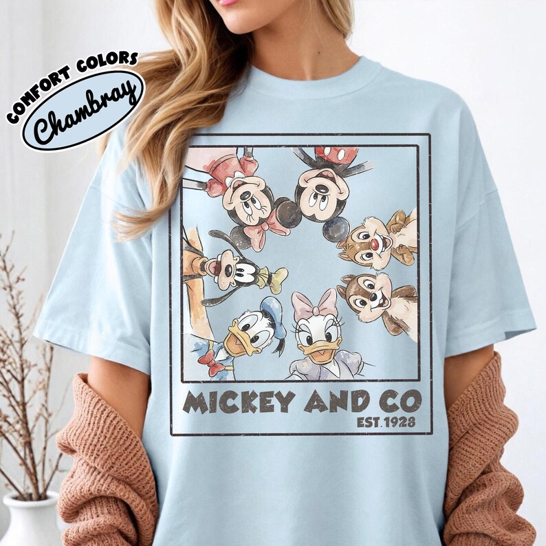 Puede incluir: Camiseta azul claro con una ilustraci&oacute;n enmarcada de Mickey Mouse, Minnie Mouse, Donald Duck, Daisy Duck, Goofy y Chip y Chop. La camiseta tiene el texto "Mickey and Co. Est. 1928" y "Comfort Colors Chambray".