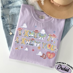 May include: A light purple t-shirt featuring the text I KNEW IT, I KNEW YOU in a stylized font filled with patterns of cowboy hats, boots, and space ranger suits. The design includes small rocket ships, stars, clouds, and a red heart with TS.