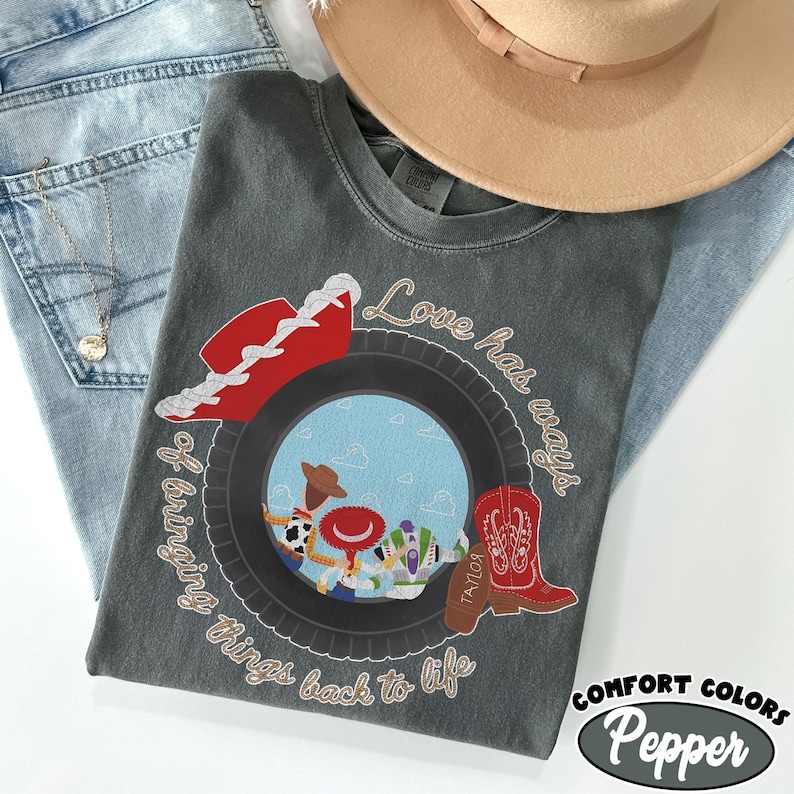 May include: A charcoal gray t-shirt featuring a graphic of a black tire framing a blue sky with clouds. Inside the tire are three cartoon characters. A red cowboy hat, a red cowboy boot, and the text Love has ways of bringing things back to life.