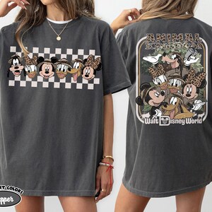 K&ouml;nnte beinhalten: Dunkelgraues T-Shirt mit einem Cartoon-Design von Disney-Figuren, darunter Micky Maus, Minnie Maus, Donald Duck und Goofy. Die Vorderseite hat ein Schachbrettmuster. Auf der R&uuml;ckseite des Shirts steht "Animal Kingdom" und "Walt Disney World".