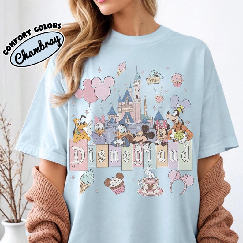May include: A light blue t-shirt featuring a graphic print of a castle, Mickey Mouse, Minnie Mouse, Donald Duck, Daisy Duck, Goofy, and Pluto. The design includes balloons, ice cream, a cupcake, a teacup, and the text Disneyland in a block font.