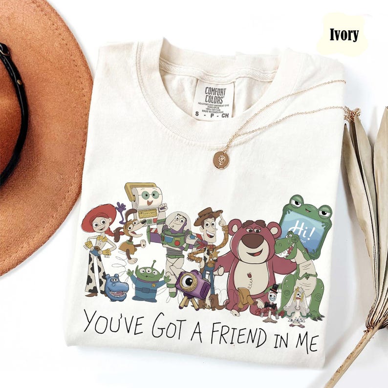 May include: An ivory short-sleeved t-shirt featuring a graphic print of various Toy Story characters including Woody, Buzz Lightyear, and Jessie. The text "YOU'VE GOT A FRIEND IN ME" is printed in black below the characters. The shirt has a crew neckline.