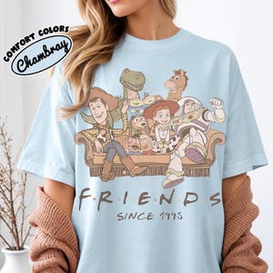 May include: Light blue t-shirt featuring a graphic of characters from the movie Toy Story, seated on a brown couch. The word "FRIENDS" is printed below the graphic, with "SINCE 1995" underneath. The shirt has the words "COMFORT COLORS" and "Chambray" printed on the top left.