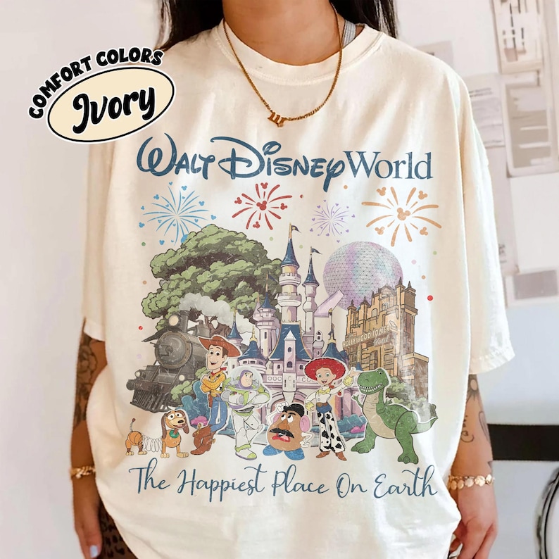 May include: Ivory-colored t-shirt featuring a colorful graphic of Walt Disney World with fireworks and cartoon characters. The text "Walt Disney World" and "The Happiest Place On Earth" are printed on the shirt.