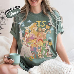 May include: A moss green t-shirt featuring a graphic print of multiple animated characters dressed in various costumes, including a space ranger, a cowgirl, and a sheriff. The text "TS" appears in yellow at the top, and a "Comfort Colors Moss" label is visible.